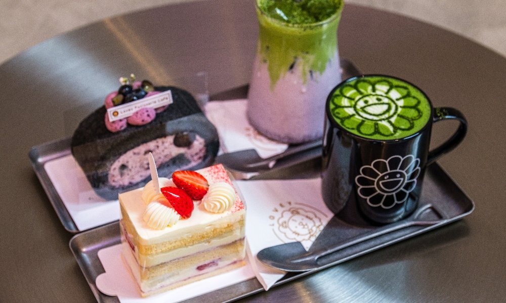 Two trays of small cakes and matcha drinks on a stainless steel table.