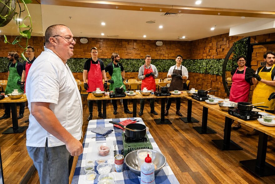 Chef standing in front of a workbench while workshop participants look on, standing behind a U-shaped arrangement of workbenches.
