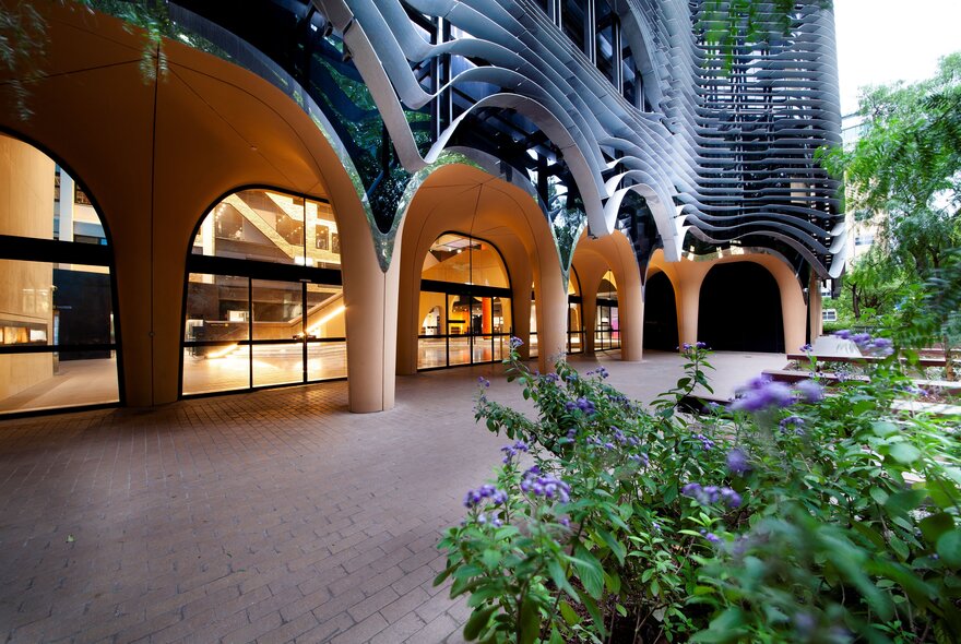 A modern building featuring a ground-floor arcade with large, smooth, light-colored concrete arches and glass entryways, contrasted by a dark, wavy, organic-shaped facade on the upper levels, with green plants and purple flowers in the foreground.