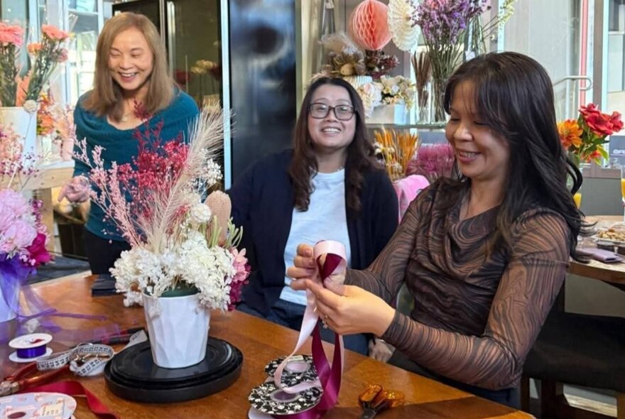 A florist working on a bow for a floral arrangement as two smiling woman watch. 