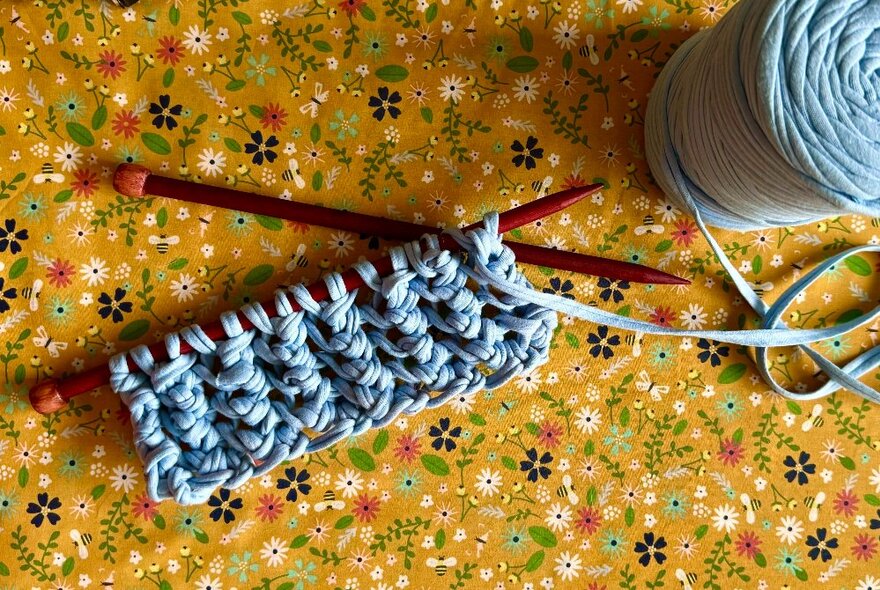 A small portion of knitting, in a blue yarn, with dark red knitting needles, displayed next to a ball of yarn and on a floral surface.