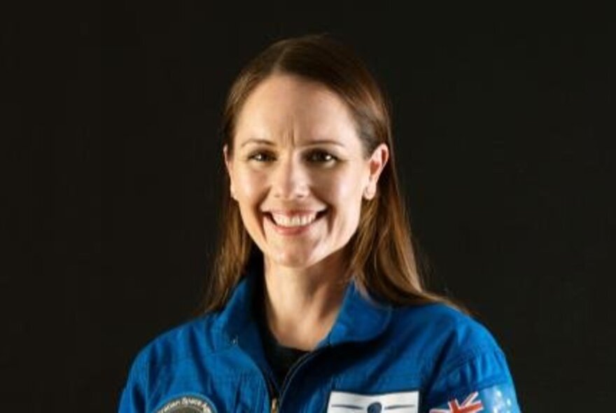 Smiling astronaut Katherine Bennell-Pegg, wearing a blue spacesuit, with her straight brown hair falling on either side of her face.
