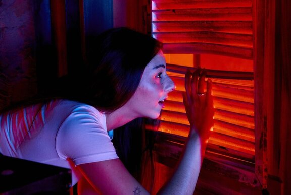 A young woman crouching to look through venetian blinds that are backlit with a red light.