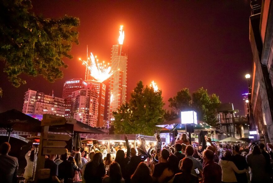 Fireworks lighting up the night sky from towers with crowds celebrating in the foreground under trees in a beer garden.