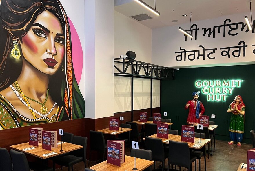 The interior of an Indian restaurant with a mural of an Indian woman on the wall, wooden tables and chairs with menu books standing on top. 