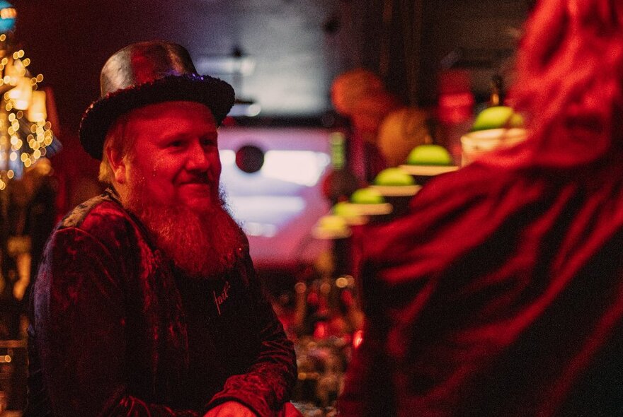 A man with a long beard and top hat in a dim bar setting  with red light cast on him. 