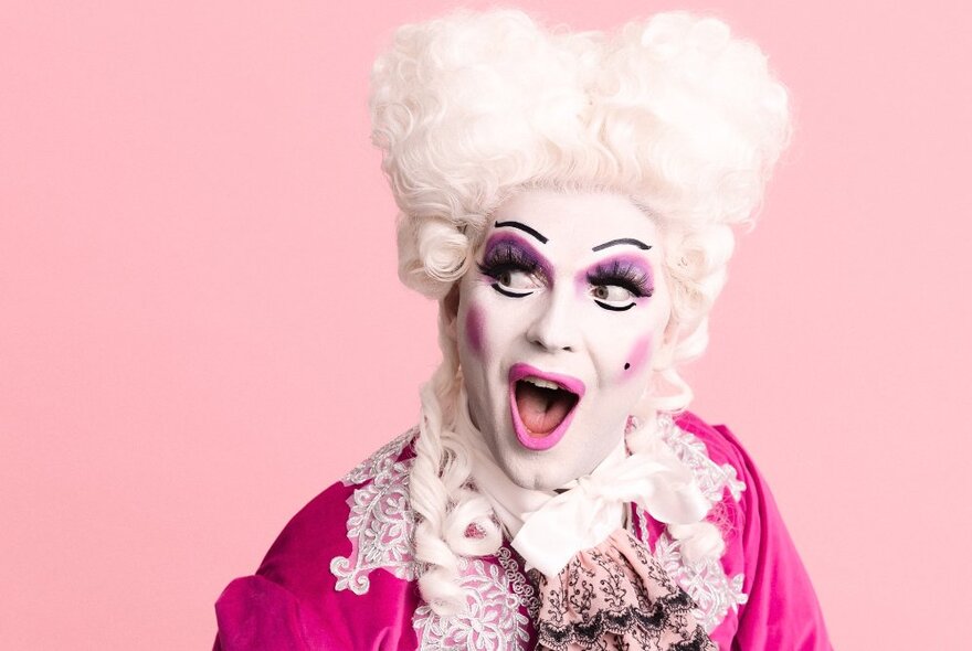 Drag queen and comedian, Adore Handel in heavy white and pink make-up with a white wig and pink ruffled clothing, with a pink background.