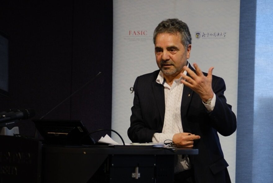 Professor Lo Bianco speaking at a lectern, gesticulating with one hand.