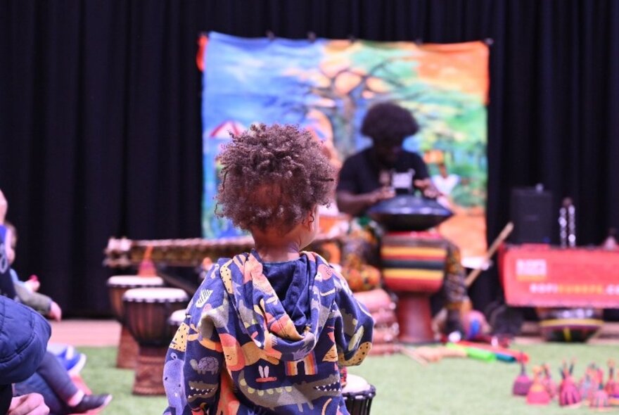 The back of a child in a room with other kids and an adult playing an African drum. 