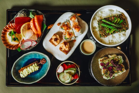 A Japanese bento tray with a selection of gourmet tasters including nori rolls, yakitori and scallops.