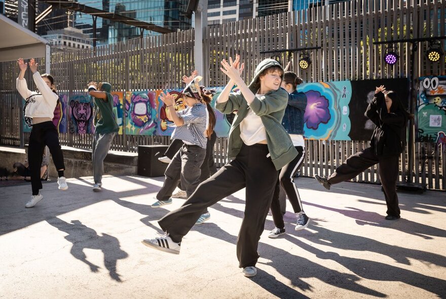 A group of hip hop dancers dancing in the sun outside, with a tall timber fence behind them with a colourful painted mural attached.
