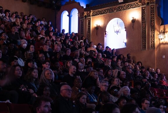 People seated in an old cinema with ornate decor. 