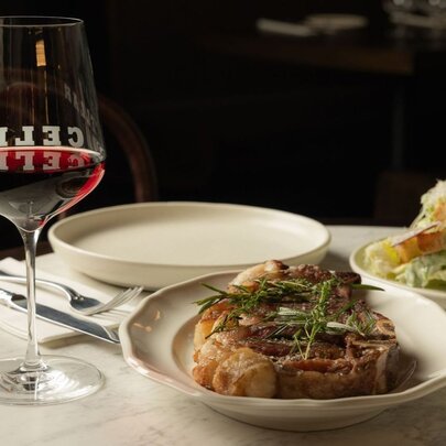 A plate with perfectly cooked bone-in steak alongside a glass of red wine on a dining table.