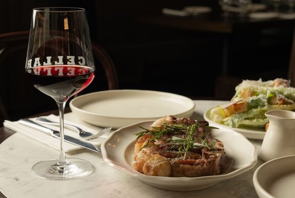 A plate with perfectly cooked bone-in steak alongside a glass of red wine on a dining table.
