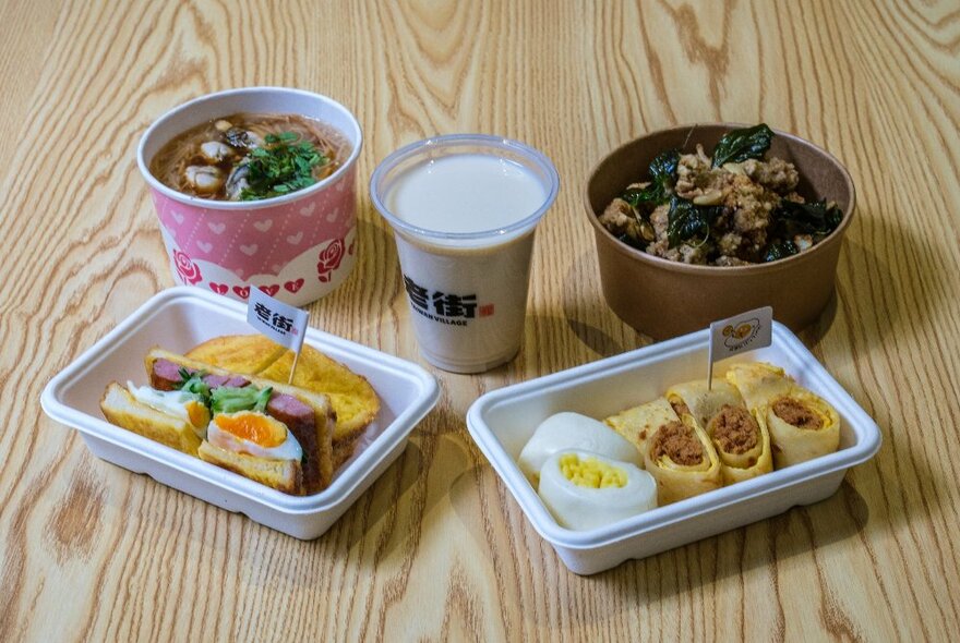 A selection of takeaway food containers from Taiwan Village includes breakfast pancakes with egg and sausage, a milky drink, and some braised meat and noodle soups.