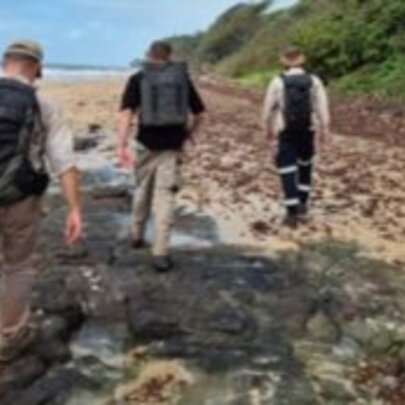 Three people with backpacks walk along a rocky shoreline next to a beach.