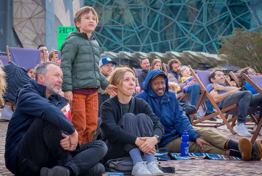 Adults and children sitting on the ground or in deckchairs, in the open air plaza at Fed Square, watching movies on the big digital screen (out of sight).