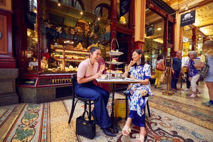 Two people seated at a cafe table with tiered cakestand in the Block Arcade outside Tea Rooms, people queuing nearby.