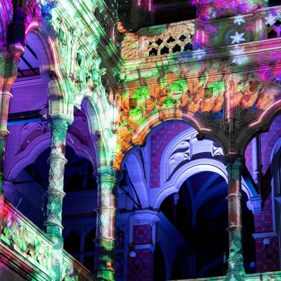 Interior view of a historic building featuring stone arches and columns, transformed by a vibrant, multi-colored light projection show.