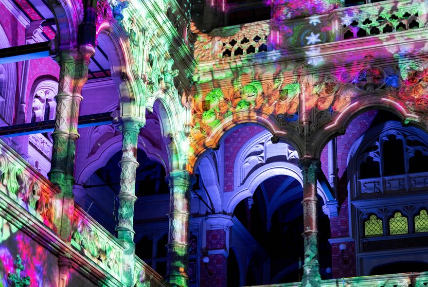 Interior view of a historic building featuring stone arches and columns, transformed by a vibrant, multi-colored light projection show.