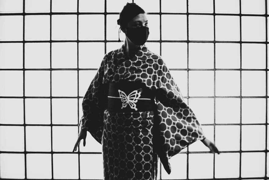A black and white image of a woman wearing a kimono with a butterfly on the obi, and a face mask.