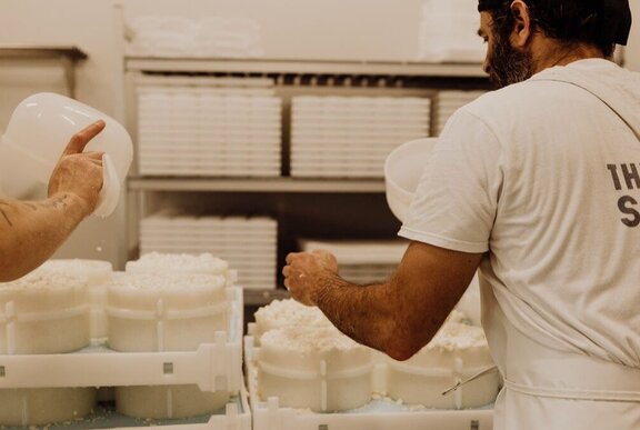 A cheesemaker dressed in white, seen from behind tending to large wheels of cheese with white rinds.