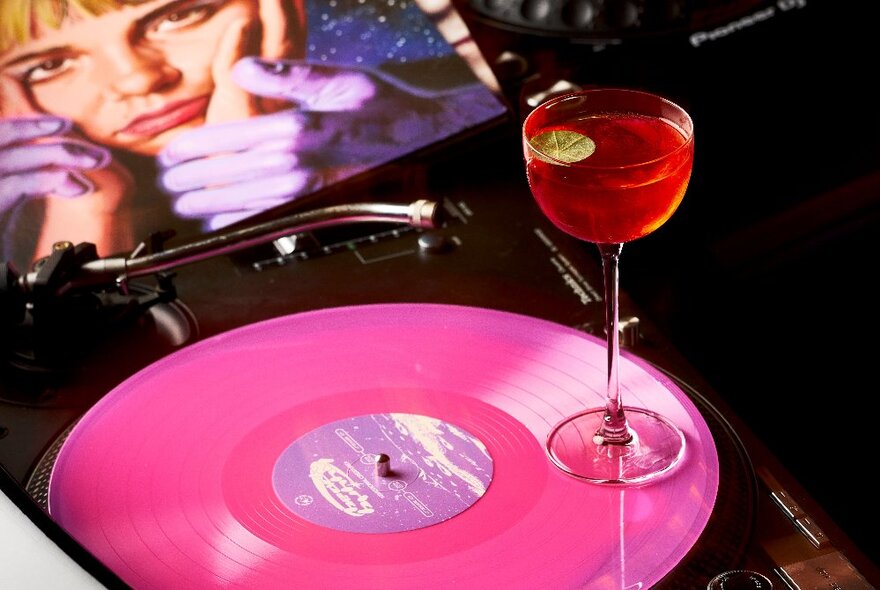 A red cocktail in a small martini glass, balanced on a pink vinyl record that rests on a turntable at Curious bar.