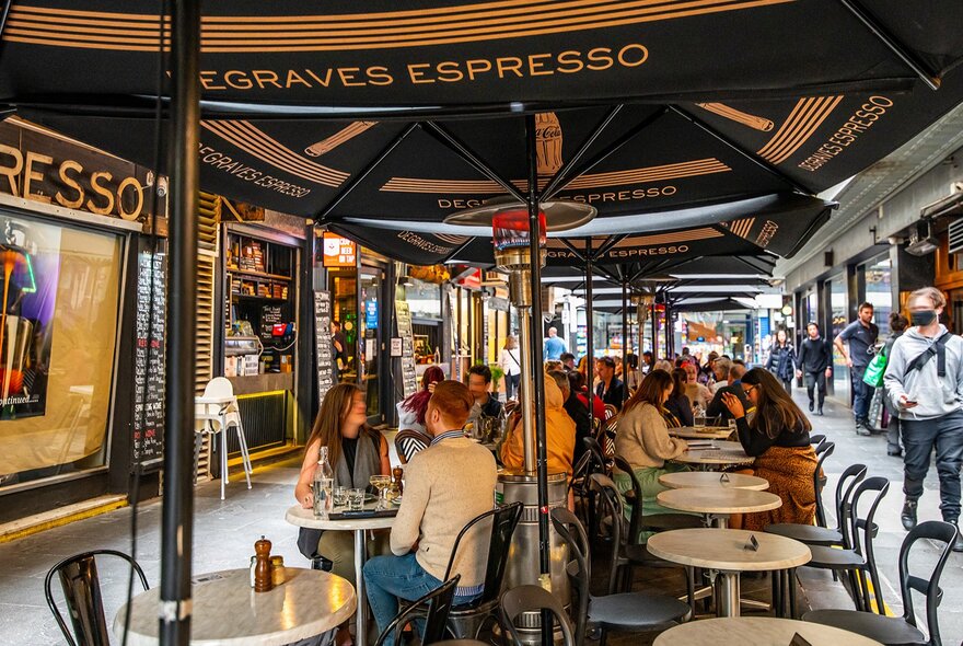 People dining at small cafe tables under umbrellas in a laneway.