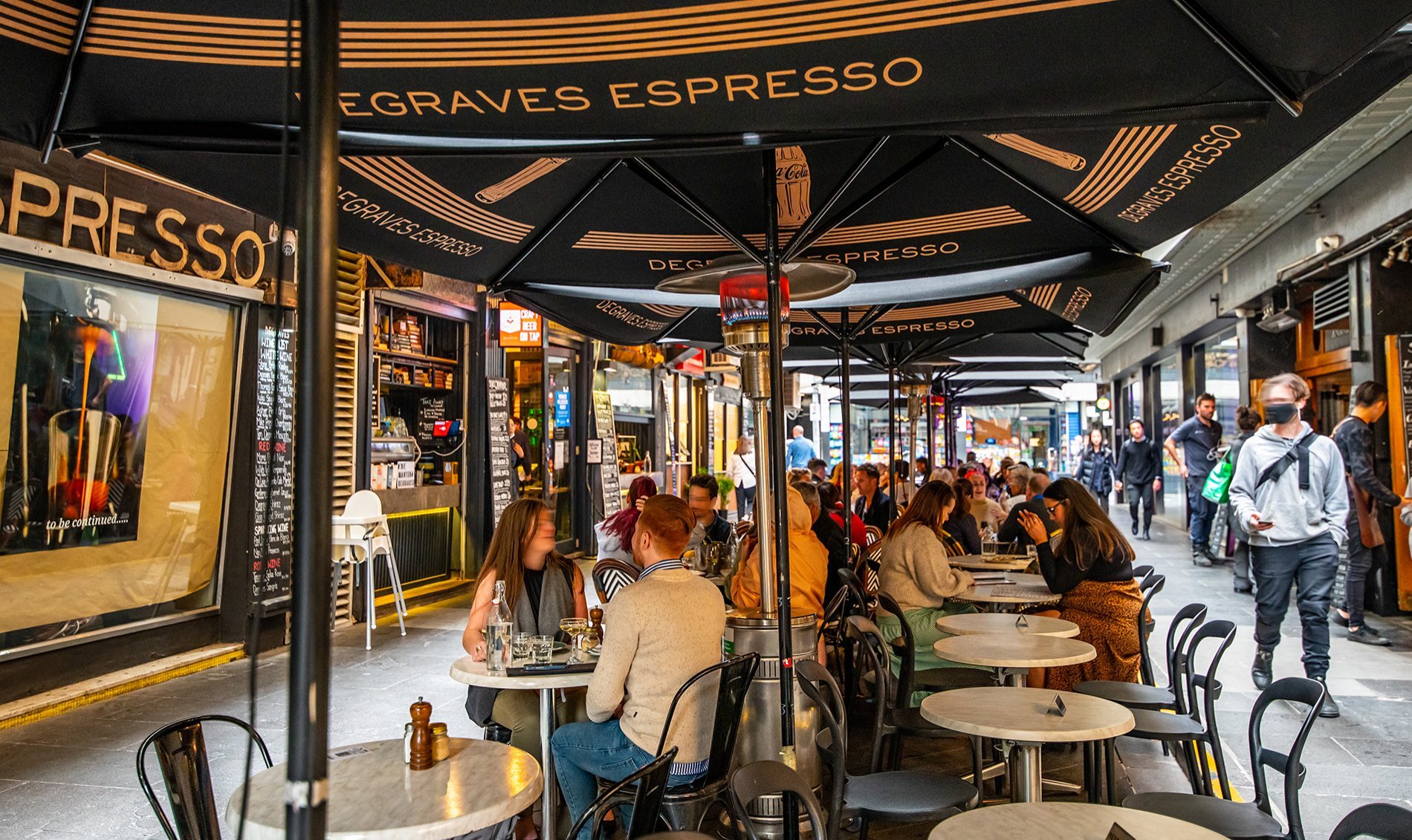 People dining at small cafe tables under umbrellas in a laneway.