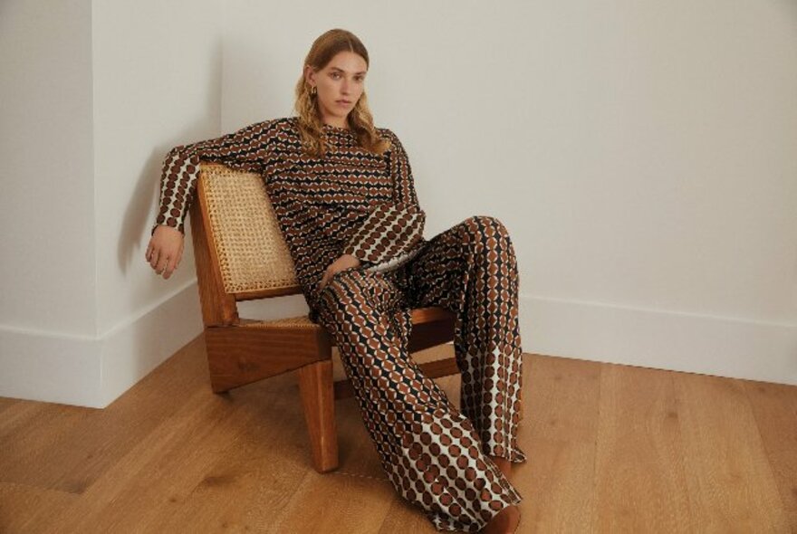 Woman with dark blonde hair lounging in low wooden chair in brown, white and black silky, loose jumpsuit.