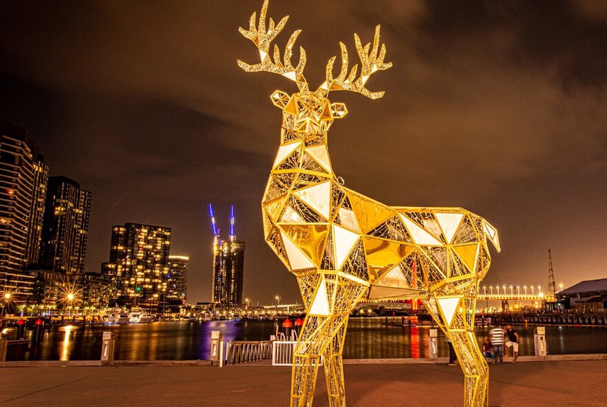 A large golden reindeer statue beside the water in a city at night. 