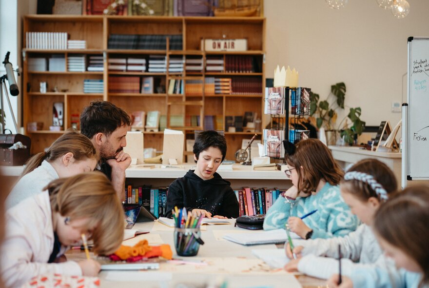 Young teens in a classroom setting with a teacher looking on and bookshelves in the background.