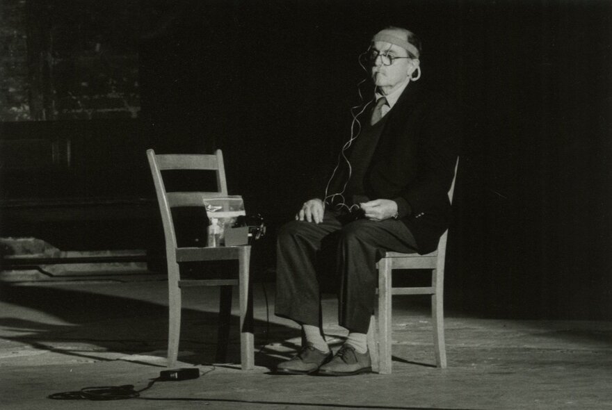 An older man wearing a suit and cap sitting on a wooden chair, facing another wooden chair with an object on it, electrical wires from his cap attached to the object, in a dimly lit space.