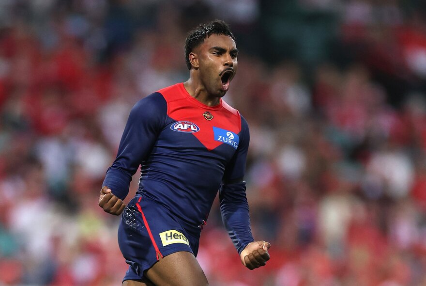 A Melbourne AFL player cheering himself and his team on the field with his mouth wide open, both his fists clenched and by his side as he runs. 