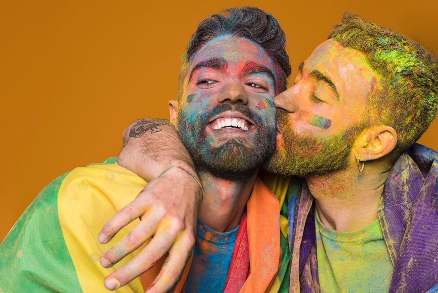 Two people covered in coloured powder, one kissing the other on the cheek, with a rainbow flag draped over the shoulders of one of them.