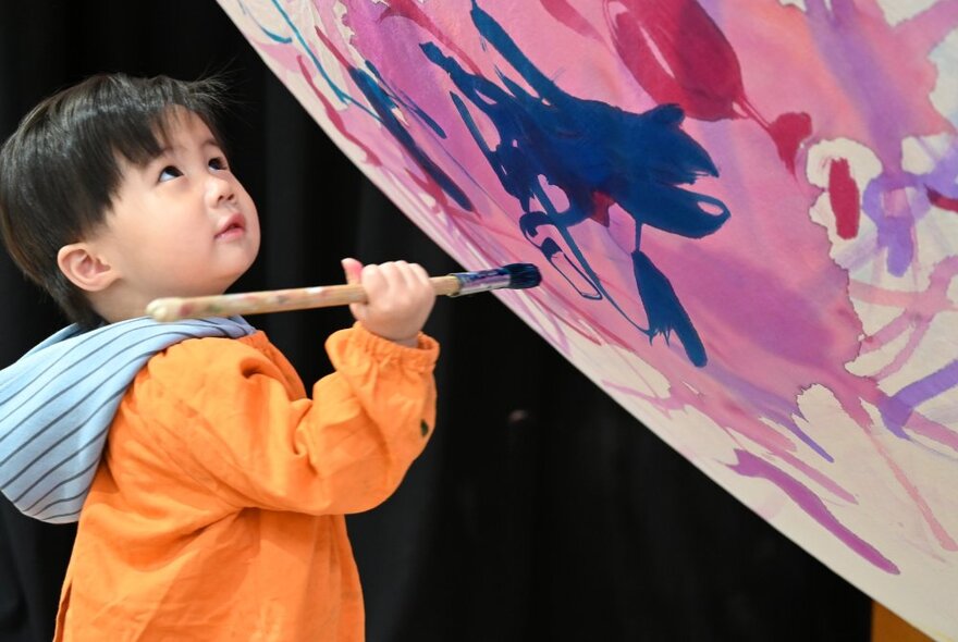 A young child in an orange smock gripping a paintbrush and daubing a wall with blue paint, the canvas is on a diagonal to the child. 