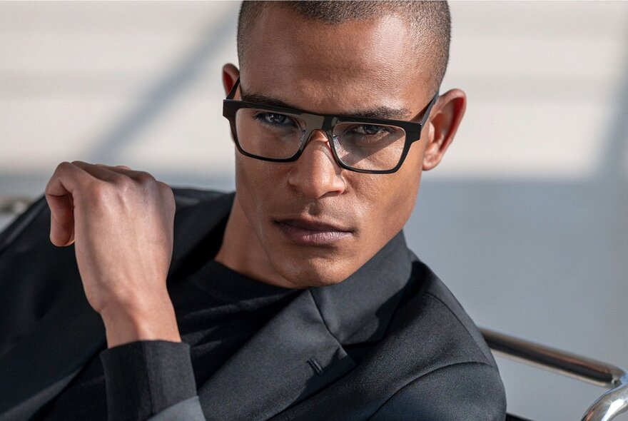 An eyewear advertisement with a man in a black suit and black eyeglasses, looking directly at the camera.