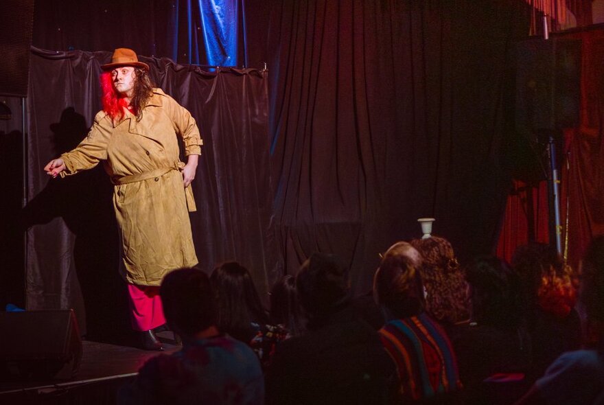 Comedian with dyed hair performing on stage, wearing a hat and trenchcoat.