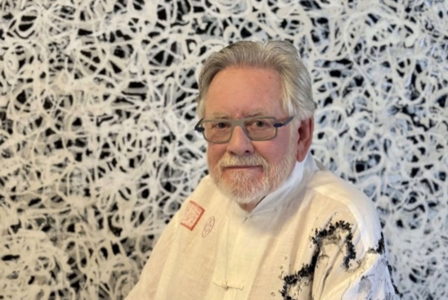 An older man in glasses and a white shirt with black paint splatters, seated in front of an artwork of white swirls on a black background.