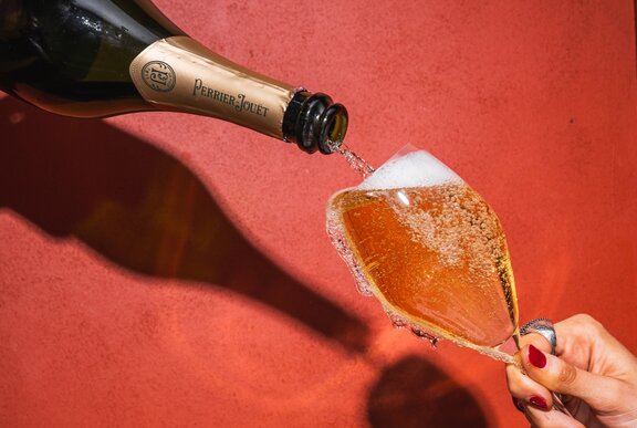 A bottle of champagne being poured into an overflowing champagne glass held on an angle by a woman's hand.