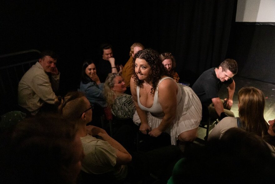 A woman in a white dress actively interacts with a seated audience in a small comedy venue, squatting on a chair directly in front of an audience member.