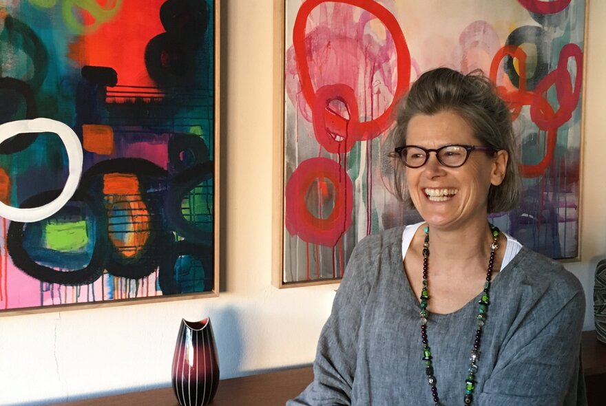 A smiling woman posed in front of two abstract artworks hanging on a wall.