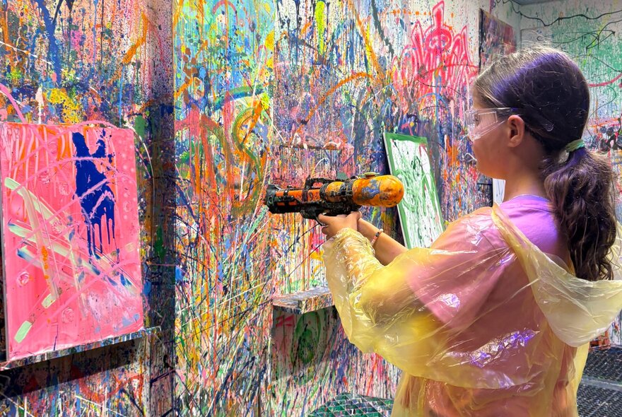 A child wearing a plastic coat, squirting paint with a paintgun at a pink canvas on a wall splattered with paint.