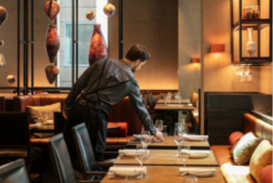 Waiter placing napkins next to plates on a dining table inside a restaurant.