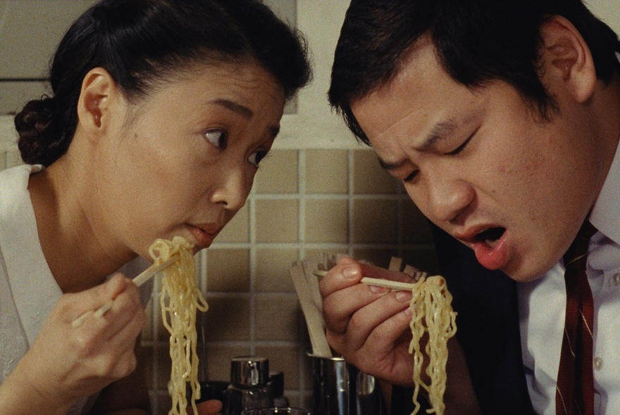 A Japanese woman and man both leaning in over the noodles they're holding in chopsticks.