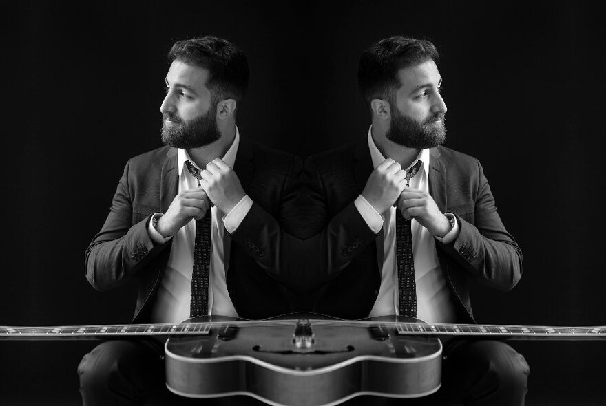 Black and white mirrored image of the guitarist Adam Russo in a suit, straightening his tie, looking to the side, a guitar resting on his lap.