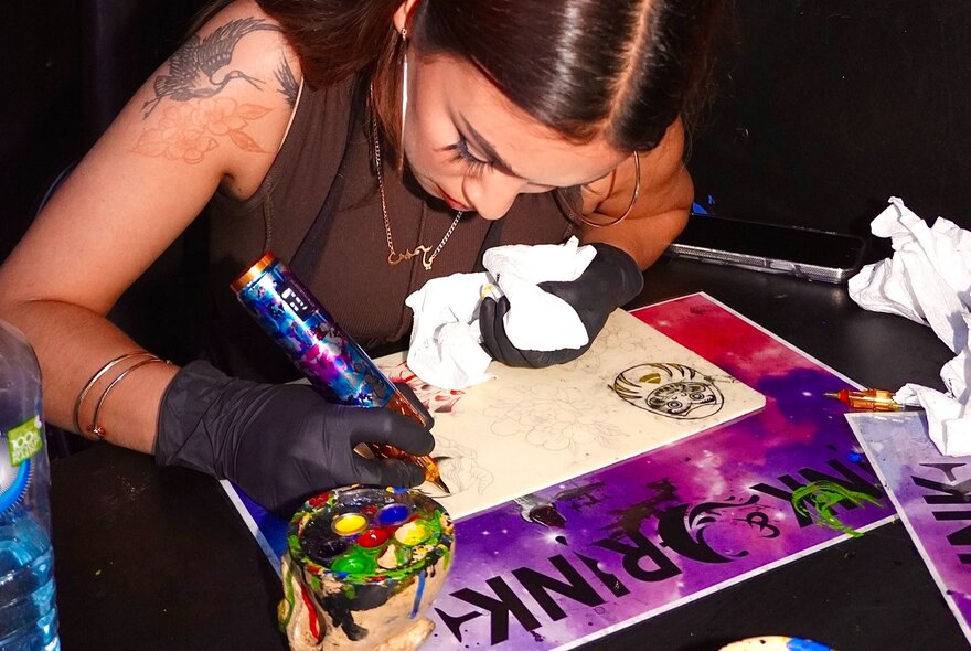 A person seated at a table, wearing black latex gloves, concentrating while using a tattoo gun to create a tattoo design on a piece of paper.