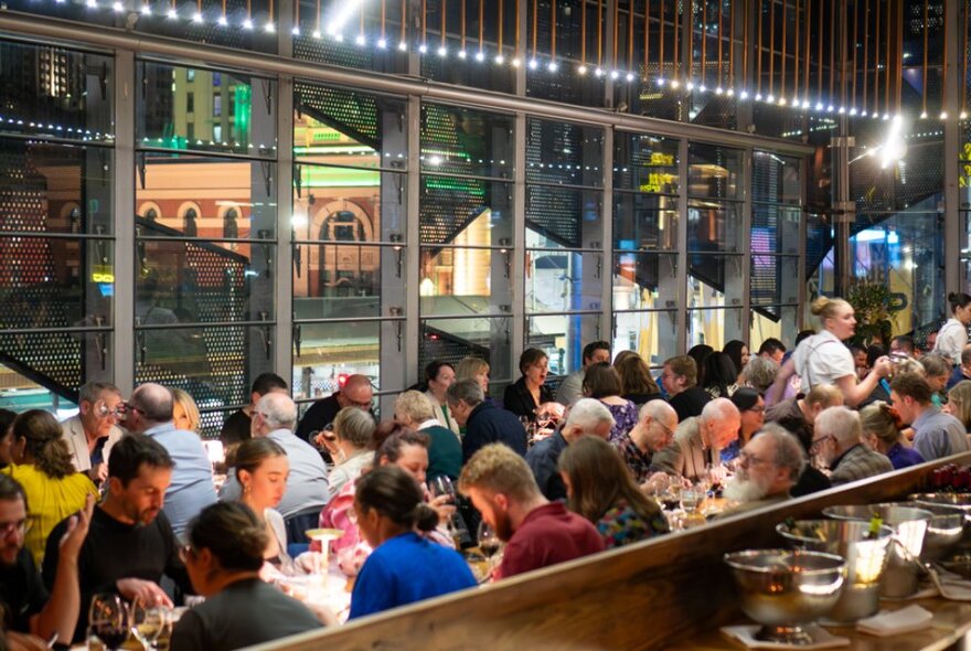 Dining patrons eating and drinking, seated at long tables inside Taxi Kitchen restaurant, with large windows surrounding them and views of Flinders Street Station facade.