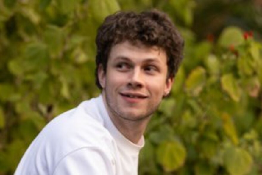 Man wearing a white skivvy, looking to the side in a garden setting.