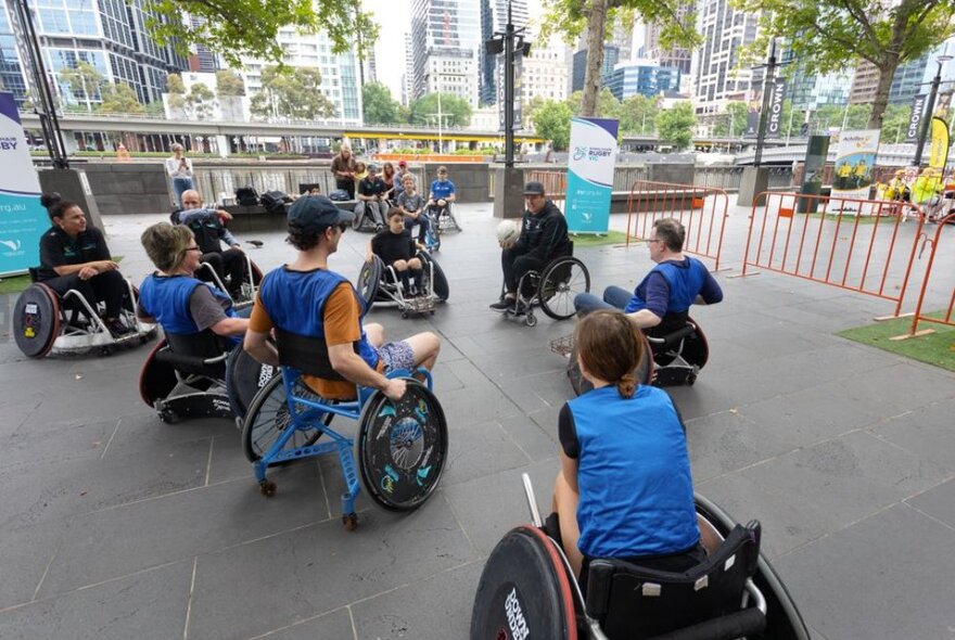 A group of people in wheelchairs playing a ball game in an open air plaza, with city views in the background.